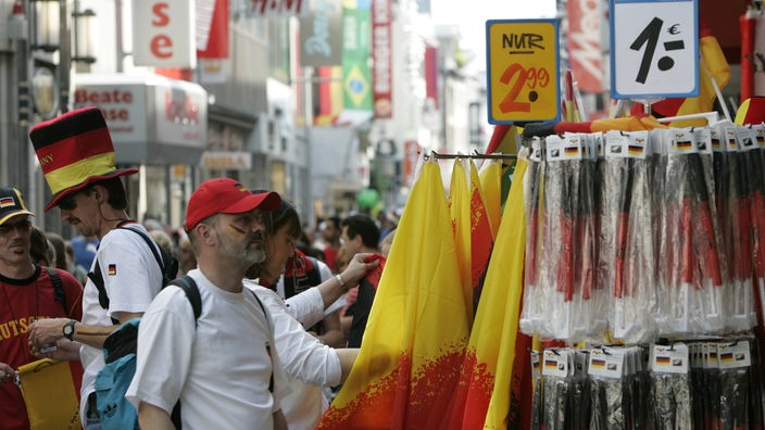 Ein Fußballfan mit roter Mütze steht an einem Verkaufsständer mit Deutschlandfahnen. Dahinter Blick in eine Kölner Einkaufsstraße, auf der sich Fußballfans drängen.