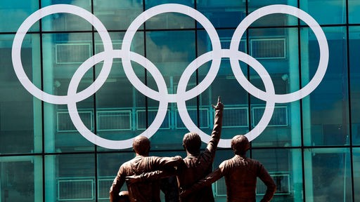 Die Olympischen Ringe am Old-Trafford-Stadion, davor ist ein Denkmal mit drei Fußballern zu sehen