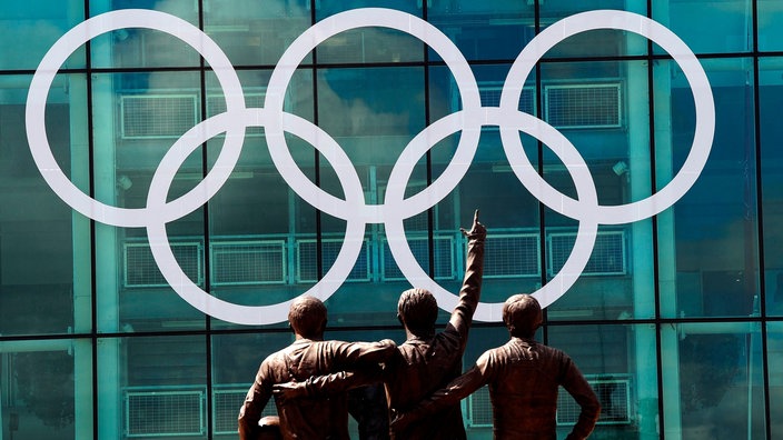 Die Olympischen Ringe am Old-Trafford-Stadion, davor ist ein Denkmal mit drei Fußballern zu sehen | Bildquelle: picture alliance / empics Die Olympischen Ringe am Old-Trafford-Stadion, davor ist ein Denkmal mit drei Fußballern zu sehen