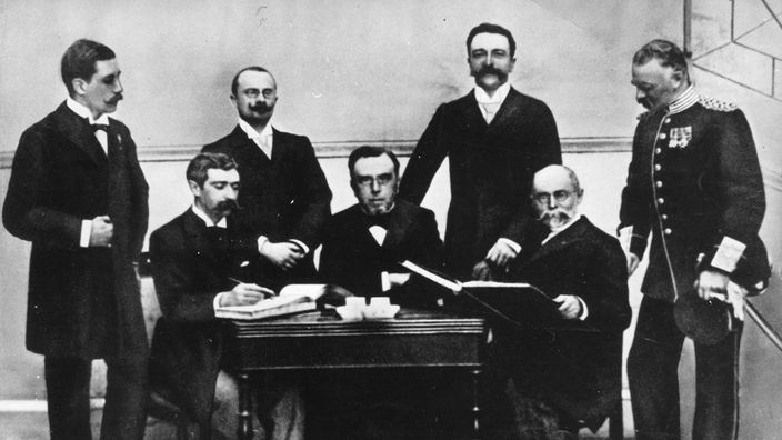 Schwarzweiß-Foto: Sieben Herren stehen oder sitzen um einen Tisch