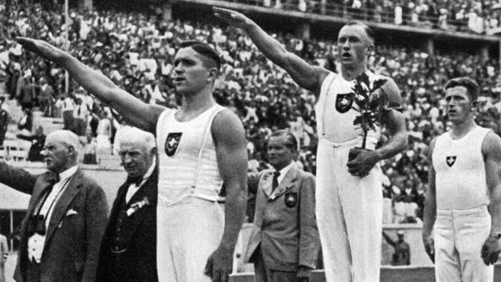 Drei Männer auf dem Gewinnertreppchen bei Olympia, zwei heben die Hand zum Hitlergruß