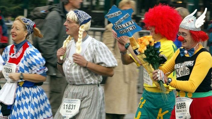 In bunten Kostümen und einem Schild mit der Aufschrift 'Bonjour Zuschauer haltet durch' laufen vier Läufer die Strecke beim Köln-Marathon 2003.
