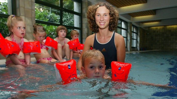 Das Bild zeigt ein Kind mit Schwimmflügeln im Wasser, das von einer jungen Frau festgehalten wird.