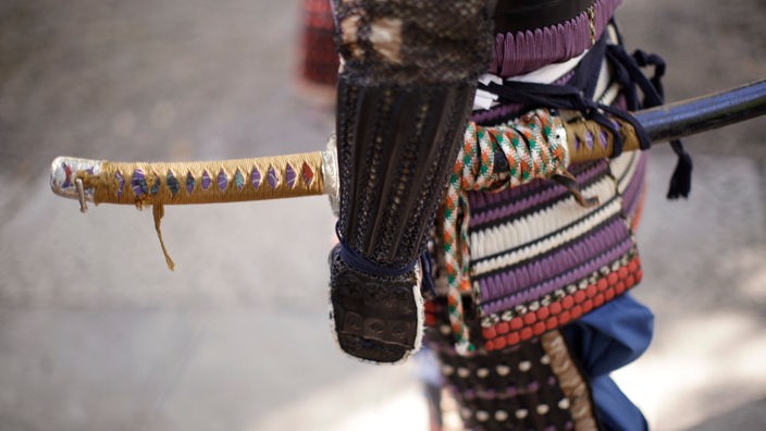 Ein Mann ist in traditioneller Samurai-Kleidung abgebildet. Prominent ist ein langes Schwert an seinem Gürtel zu sehen
