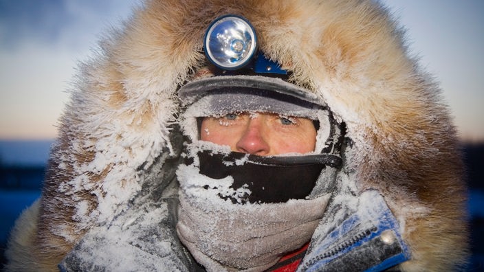 Gesicht von Extremsportler in eisiger Kälte mit gefrorenen Wimpern, eingepackt in Felljacke mit dickem Schal. Nur die Augen bleiben frei.