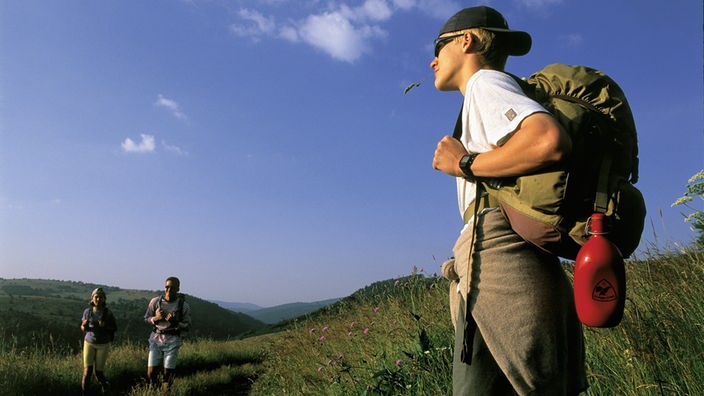Ein junger Wanderer steht am Wegesrand. Im Hintergrund sind zwei weitere junge Wanderer zu erkennen.