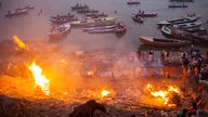 Mehrere Menschen stehen um ein großes Feuer an einem Fluss, im Hintergrund treiben Boote