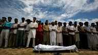 Eine muslimische Trauergemeinde steht vor einem in ein Tuch gewickelten Leichnam unter freiem Himmel