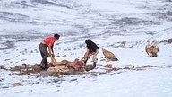 Tibetische Domdens, Knochebrecher, bereiten eine Leiche auf einer verschneiten Ebene für die Luftbestattung vor, um sie herum sitzen Geier