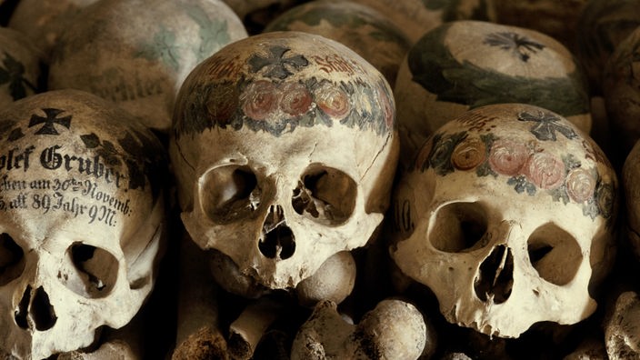 Gestapelte Knochen und Totenschädel in einem Beinhaus in Österreich. Die Schädel sind kunstvoll mit einem Blumenmuster bemalt. Außerdem tragen sie den Namen der Verstorbenen und das Todesdatum.