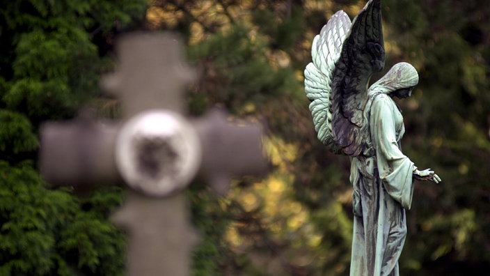Vor grünen Bäumen und Sträuchern erkennt man schemenhaft die Umrisse eines Grabkreuzes. Rechts daneben die grün oxydierte Metallstatue eines beflügelten Engels mit gesenktem Haupt. | Bildquelle: ddp Vor grünen Bäumen und Sträuchern erkennt man schemenhaft die Umrisse eines Grabkreuzes. Rechts daneben die grün oxydierte Metallstatue eines beflügelten Engels mit gesenktem Haupt.