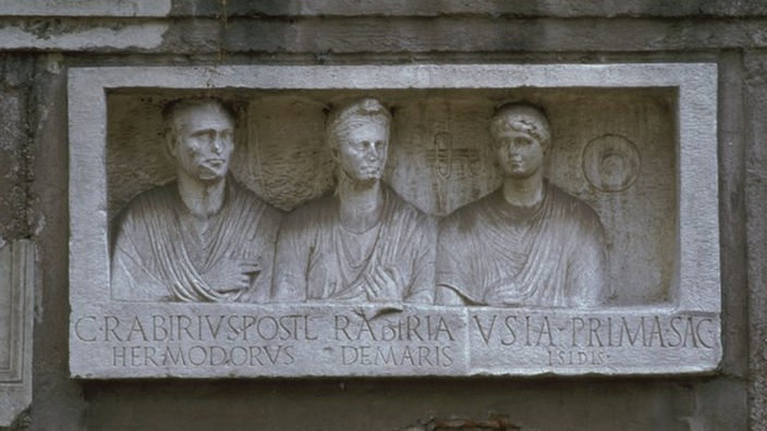 Ein römisches Grabmal im Querformat zeigt im Relief die Abbilder von drei Verstorbenen. Unter den Reliefs sind die Namen der drei in Stein gemeißelt.