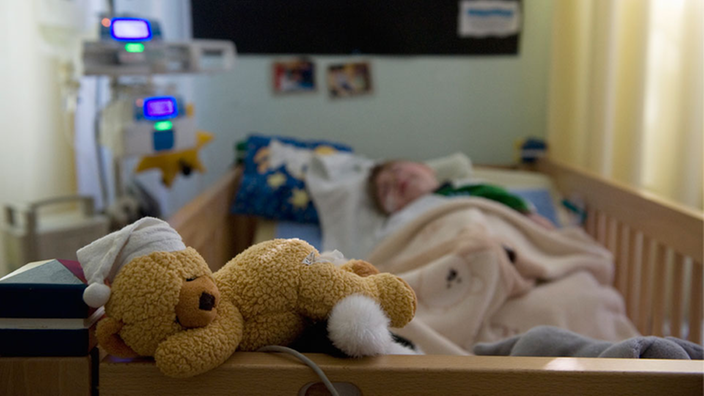 Ein Kind im Krankenbett. Am Fußende liegt ein Teddy