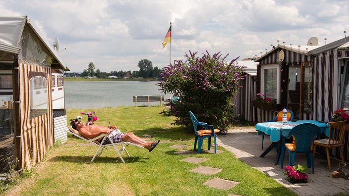Mann sonnt sich auf einem Campingplatz vor einem bunt gestreiften Zelt auf einem Campingstuhl. | Bildquelle: dpa/Sebastian Gollnow Mann sonnt sich auf einem Campingplatz vor einem bunt gestreiften Zelt auf einem Campingstuhl.