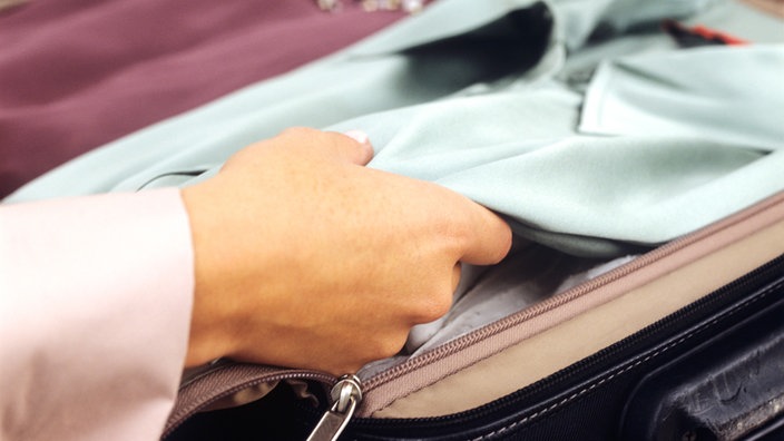 Eine Frau legt eine gefaltete Bluse in einen Koffer. | Bildquelle: Picture alliance Eine Frau legt eine gefaltete Bluse in einen Koffer.