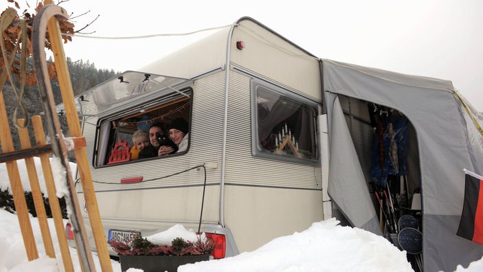 Das Foto zeigt einen Wohnwagen mit Vorzelt im Tiefschnee. Aus dem hinteren Fenster schauen mehrere Menschen heraus und lächeln.