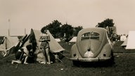 Das Schwarzweiß-Foto zeigt zwei junge Frauen auf der Wiese vor einem Zelt. Daneben steht ein VW Käfer.