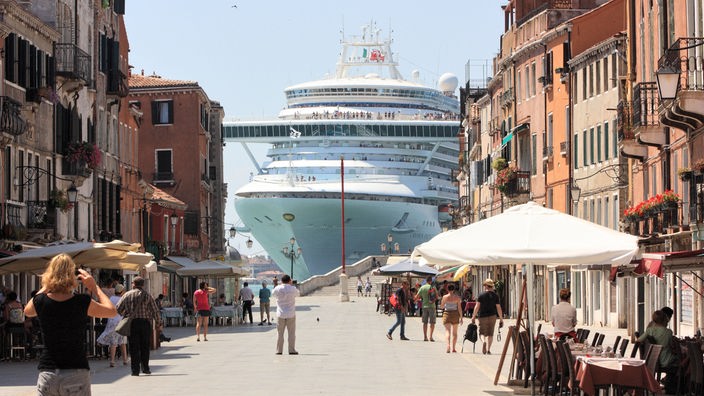 Eine Straße mit Cafés in der Innenstadt von Venedig, im Hintergrund ein riesiges Kreuzfahrtschiff 