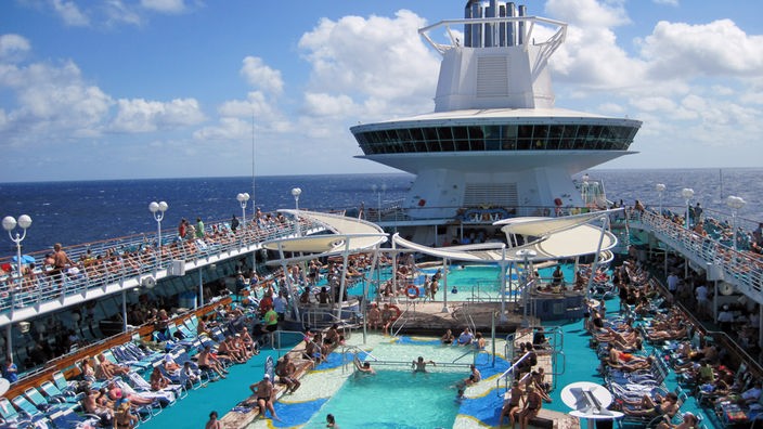 Ein Swimmingpool auf dem Deck eines Kreuzfahrtschiffes, in dem viele Passagiere baden. Im Hintergrund gibt ein Schornstein schwarzen Rauch ab.