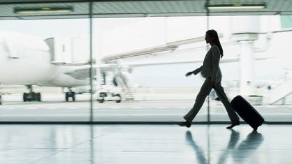 Geschäftsfrau geht mit Koffer am Flughafen. Im Hintergrund sind durch eine Scheibe Flugzeuge zu erkennen.
