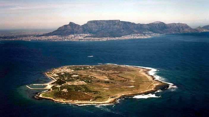 Eine grüne Insel im blauen Meer. Im nördlichen, hinteren Bereich ist der Gebäudekomplex der Haftanstalt. Im Hintergrund die Küste mit Kapstadt.