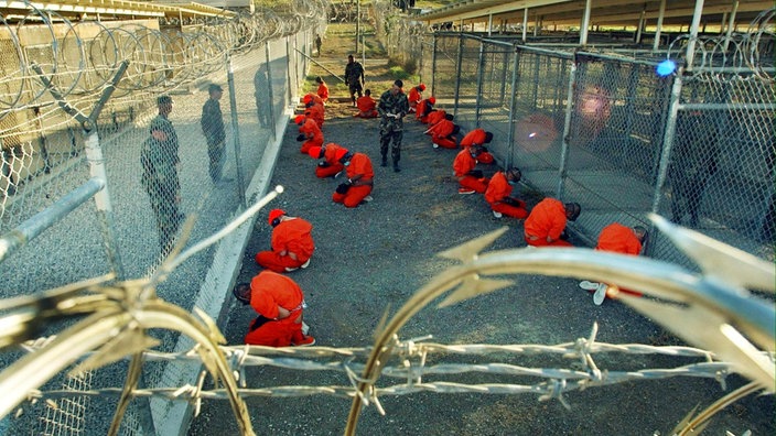 18 Gefangene in orangefarbenen Overalls knien auf einem offenen Platz, hoch mit Stacheldraht umzäunt, daneben fünf bewaffnete US-Militärpolizisten. | Bildquelle: picture-alliance/ dpa / DB Shane T. Mccoy 18 Gefangene in orangefarbenen Overalls knien auf einem offenen Platz, hoch mit Stacheldraht umzäunt, daneben fünf bewaffnete US-Militärpolizisten.