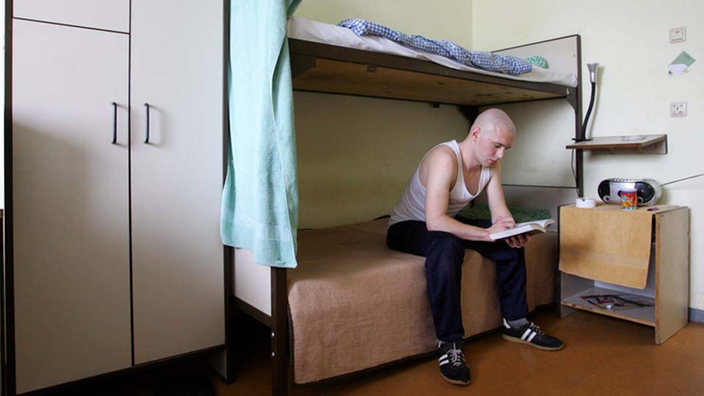Ein Häftling sitzt auf seinem Bett in seiner Zelle. Er liest in einem Buch.