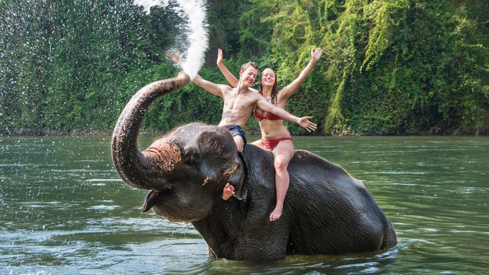 Zwei Jugendliche sitzen auf Elefanten und werden mit dem Rüssel mit Wasser nass gespritzt