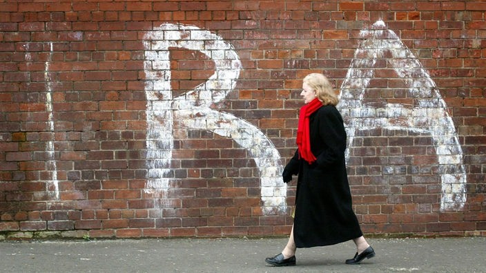 Eine Frau geht vor einer Mauer lang, auf der IRA steht