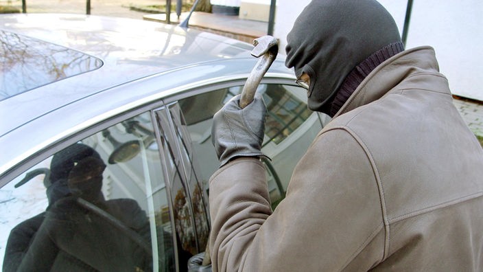 Maskierter Einbrecher bricht mit einem Brecheisen ein Auto auf.