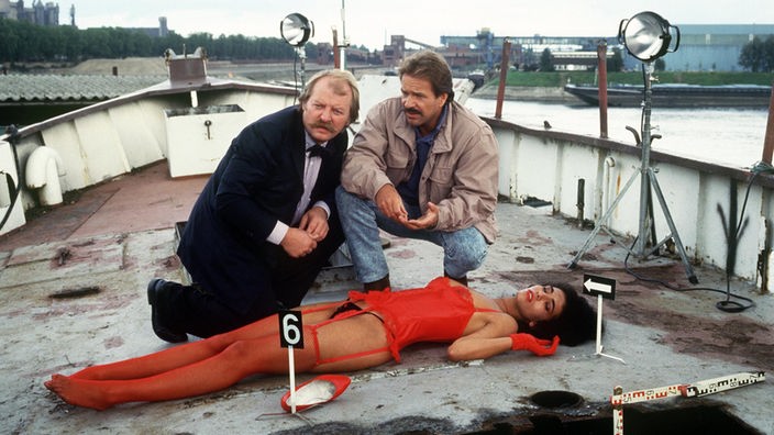 Zwei Männer und eine Tote auf einem Boot. Männer knien beide, vor ihnen tote Prostituierte in roter Reizunterwäsche.
