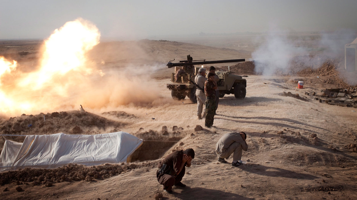 Ein Artilleriefahrzeug steht auf einer Düne in der irakischen Wüste und feuert, während sich einige Kämpfer schützend zu Boden ducken.