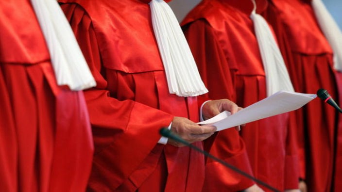 Nahaufnahme von vier roten Roben der Bundesverfassungsrichter; einer hält ein Blatt Papier vor sich.