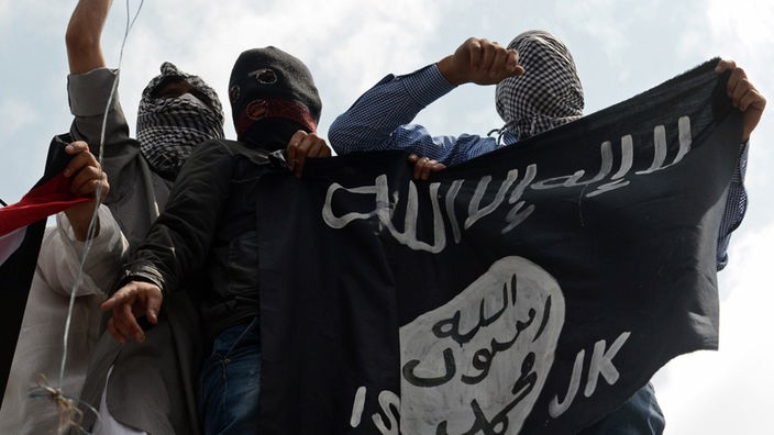 Vermummte Demonstranten halten eine Flagge des IS hoch.