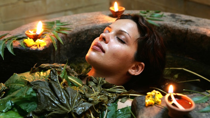 Eine Frau liegt mit geschlossenen Augen in einer steinernen Wanne. Sie ist bis zum Hals mit Blättern bedeckt. Um sie herum stehen Kerzen.