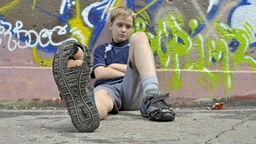 Junge sitzt mit kaputten Schuhen und traurigem Blick vor einer Mauer.