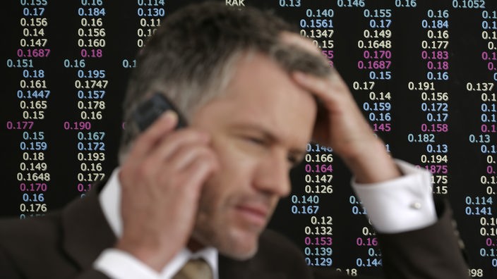 Börsenmakler telefoniert mit seinem Handy vor der Anzeigetafel für die Börsenkurse