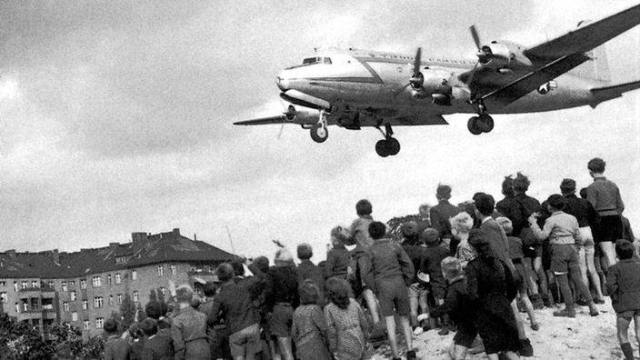 Die Schwarz-Weißfotografie zeigt einen sogenannten Rosinenbomber nach dem Zweiten Weltkrieg in Berlin, der von einer Menschenmenge erwartet wird.