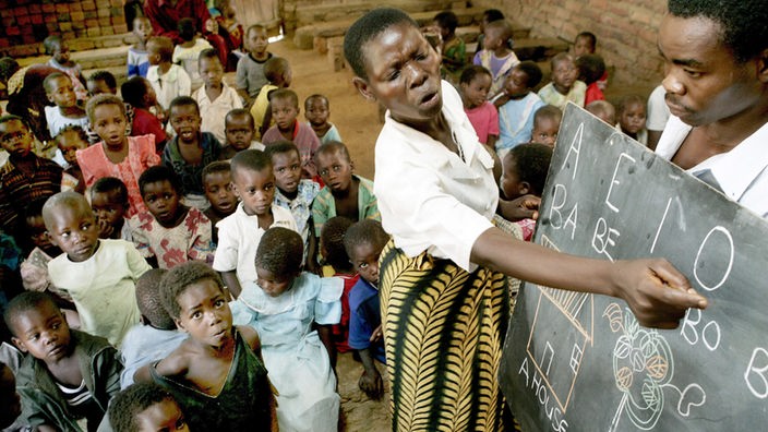Eine Lehrerin unterrichtet eine große Gruppe kleiner Kinder in einer Schule ohne Tische oder Bänke.