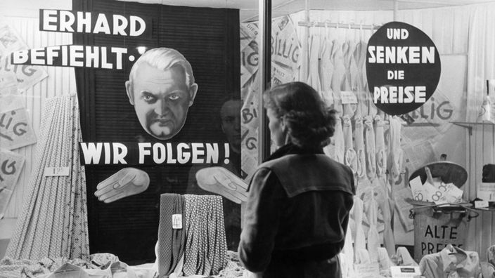 Eine Frau vor einer Schaufenster-Auslage. Im Schaufenster ein Plakat mit der Aufschrift "Erhard befiehlt – wir folgen ...und senken die Preise"