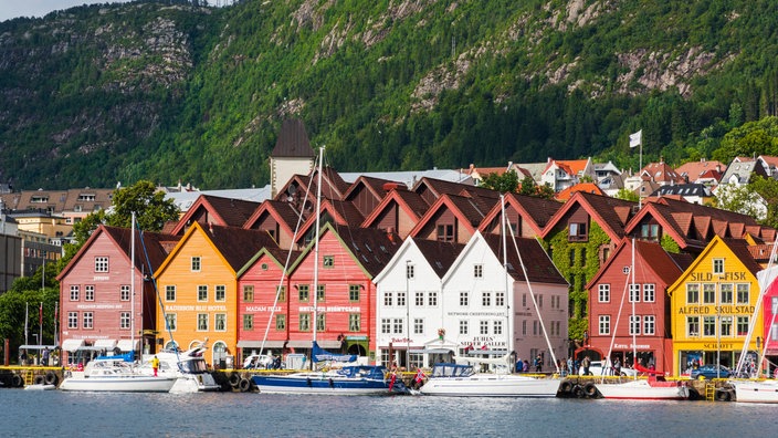 Vor einer Bergkulisse stehen kleine Häuser im Hafen von Bergen, Norwegen | Bildquelle: picture alliance / Westend61 Vor einer Bergkulisse stehen kleine Häuser im Hafen von Bergen, Norwegen