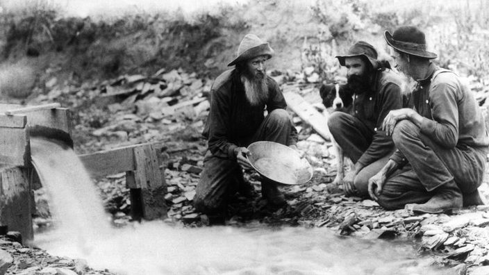 Schwarzweiß-Foto: Drei Goldgräber an einem Fluss