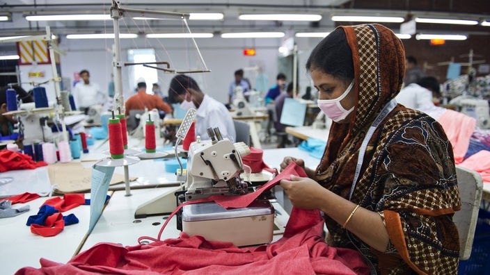 Frau mit Mundschutz sitzt in einer Fabrik an der Nähmaschine