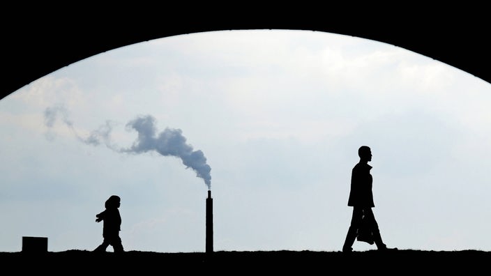 Die Silhouette zweier Passanten vor einem rauchenden Schornstein im Hintergrund