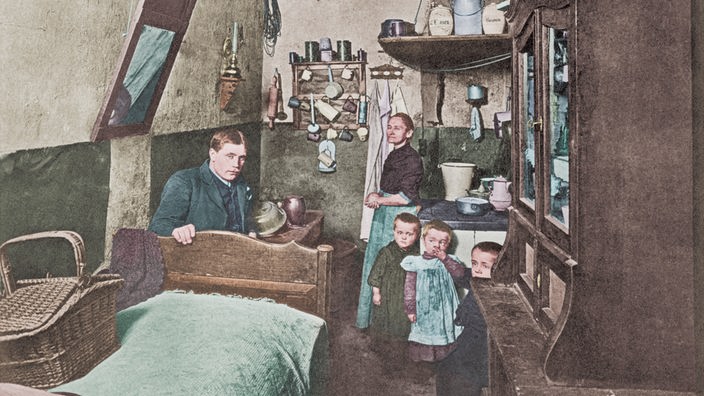 Nachkoloriertes Foto eines engen, schmalen Raumes; die Mutter der Familie steht im Hintergrund vor einem Herd mit Küchenutensilien, der Vater sitzt eingezwängt zwischen Tisch und Bett; vor der Mutter drei kleine Kinder.