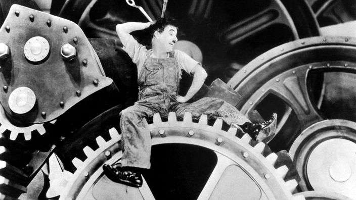 Filmausschnitt aus "Moderne Zeiten": Chaplin liegt auf einem gigantischen Zahnrad und montiert daran mit zwei Schraubenschlüsseln.