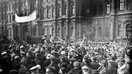 Eine Menschenmenge steht 1917 vor dem Petrograder Winterpalast. Auf einem Transparent steht auf russisch "Gebt Brot!"