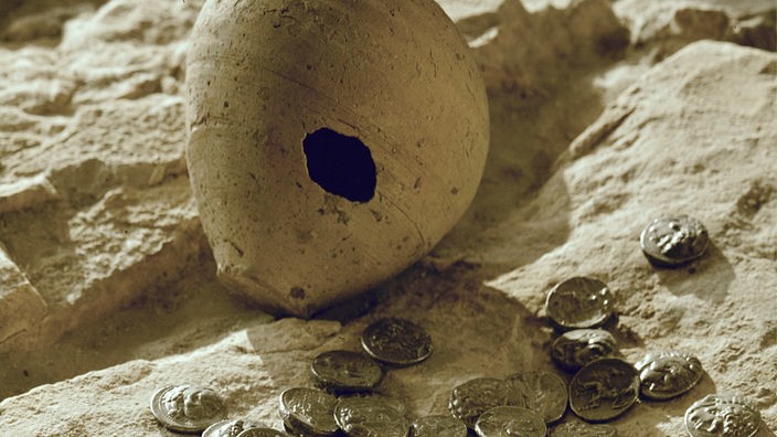 Ein Tonkrug mit einem Loch liegt auf einem Lehmboden, davor antike Münzen.
