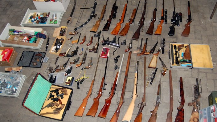 Auf dem Boden verteilt liegen zahlreiche Gewehre und Pistolen für das Bild aufgereiht.