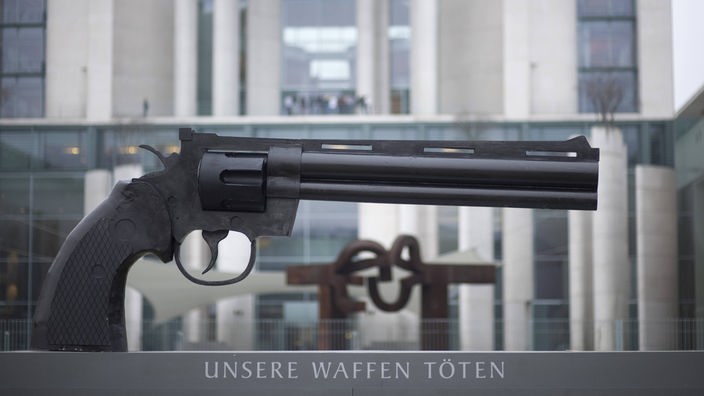 Die Skulptur eines Revolvers auf einer Steintafel mit der Aufschrift 'Unsere Waffen töten'.